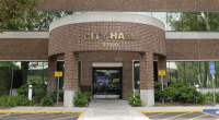 “Front entrance of a City Hall building with the address number 23920 displayed above the doorway. The structure has a red brick exterior with large glass windows. There are ‘Loading Zone’ signs on both sides of the entrance, greenery along the walkway, and a designated accessible parking space to the right.”