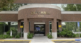 “Front entrance of a City Hall building with the address number 23920 displayed above the doorway. The structure has a red brick exterior with large glass windows. There are ‘Loading Zone’ signs on both sides of the entrance, greenery along the walkway, and a designated accessible parking space to the right.”