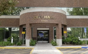 “Front entrance of a City Hall building with the address number 23920 displayed above the doorway. The structure has a red brick exterior with large glass windows. There are ‘Loading Zone’ signs on both sides of the entrance, greenery along the walkway, and a designated accessible parking space to the right.”