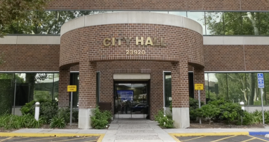 “Front entrance of a City Hall building with the address number 23920 displayed above the doorway. The structure has a red brick exterior with large glass windows. There are ‘Loading Zone’ signs on both sides of the entrance, greenery along the walkway, and a designated accessible parking space to the right.”