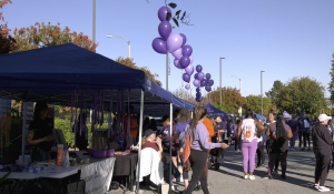 “A community event with people gathered around outdoor tents and booths on a sunny day. Purple balloons are strung in clusters along the walkway, and many attendees are wearing purple clothing. Some people are walking, while others are talking or visiting booths. Trees with autumn foliage and streetlights line the background.”