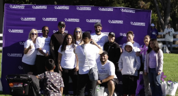 A group of people pose for a photo in front of a purple backdrop with the "Alzheimer's Association" logo repeated across it. Many of the participants are wearing white T-shirts with a message supporting the fight to end Alzheimer's. A few individuals hold colorful pinwheel flowers, a symbol commonly used in Alzheimer's awareness events. One woman in the foreground is taking a picture of the group, while a young child and stroller are also visible. The setting appears to be a sunny outdoor event in a park.