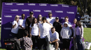 A group of people pose for a photo in front of a purple backdrop with the "Alzheimer's Association" logo repeated across it. Many of the participants are wearing white T-shirts with a message supporting the fight to end Alzheimer's. A few individuals hold colorful pinwheel flowers, a symbol commonly used in Alzheimer's awareness events. One woman in the foreground is taking a picture of the group, while a young child and stroller are also visible. The setting appears to be a sunny outdoor event in a park.