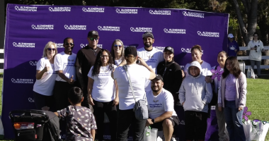 A group of people pose for a photo in front of a purple backdrop with the "Alzheimer's Association" logo repeated across it. Many of the participants are wearing white T-shirts with a message supporting the fight to end Alzheimer's. A few individuals hold colorful pinwheel flowers, a symbol commonly used in Alzheimer's awareness events. One woman in the foreground is taking a picture of the group, while a young child and stroller are also visible. The setting appears to be a sunny outdoor event in a park.