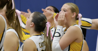 A group of female volleyball players in yellow, white, and navy uniforms cheer enthusiastically from the sidelines during a game. Most of them have their hair tied back with pink ribbons, likely in support of breast cancer awareness. One player in the foreground cups her hands around her mouth as she shouts encouragement. The players display energetic and supportive body language, emphasizing team spirit and camaraderie.