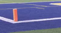 A close-up view of a football field end zone with blue turf, white boundary lines, and an orange pylon marking the corner of the goal line.