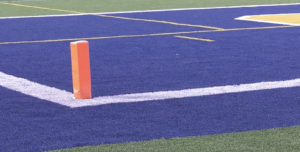 A close-up view of a football field end zone with blue turf, white boundary lines, and an orange pylon marking the corner of the goal line.