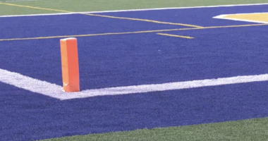 A close-up view of a football field end zone with blue turf, white boundary lines, and an orange pylon marking the corner of the goal line.