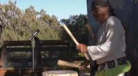 A person dressed in traditional attire plays a large wooden drum outdoors, using two drumsticks. A microphone stands nearby, and trees and a building are visible in the background under a clear sky
