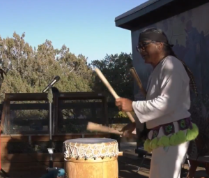 A person dressed in traditional attire plays a large wooden drum outdoors, using two drumsticks. A microphone stands nearby, and trees and a building are visible in the background under a clear sky