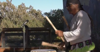 A person dressed in traditional attire plays a large wooden drum outdoors, using two drumsticks. A microphone stands nearby, and trees and a building are visible in the background under a clear sky