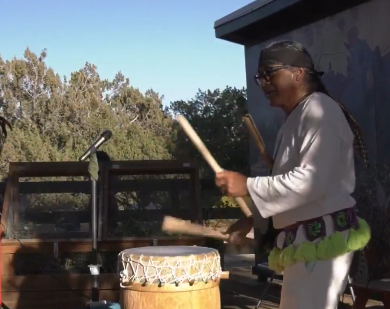 A person dressed in traditional attire plays a large wooden drum outdoors, using two drumsticks. A microphone stands nearby, and trees and a building are visible in the background under a clear sky