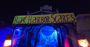 A Halloween-themed entrance display at night features a yellow sign reading “Bar Harbor Scares” in spooky black lettering. The walkway beneath is draped in black plastic and glowing blue light, with cobweb decorations and a hanging skeleton inside. Two lanterns with orange light flank the entrance, enhancing the eerie atmosphere.