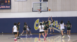 several players clustered near the basket as a ball arcs toward the hoop. The players are wearing green and white or all-white uniforms. Two players in green and white (one of them number 30) are jumping with their arms up, contesting the shot against a player in all white. Other players are scattered on the court, watching the action. A scoreboard in the upper left corner shows the score as 3 to 0. The gym wall behind the players is blue with a large yellow and white logo that starts with "Co." The court has a polished, wooden floor.