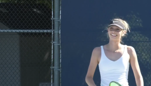 Freshman tennis player Emma Chodur’s wearing a white tank top and dark hat holding a tennis racket smiles while standing on a tennis court at college of the canyons.