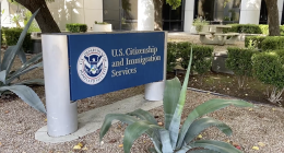 Sign for U.S. Citizenship and Immigration Services outside a building, surrounded by desert landscaping with agave plants, small bushes, and a picnic table in the background