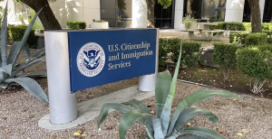 Sign for U.S. Citizenship and Immigration Services outside a building, surrounded by desert landscaping with agave plants, small bushes, and a picnic table in the background