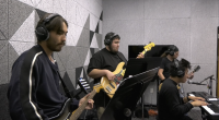 Four musicians play together in a soundproof studio. One person in the foreground plays an electric guitar, another plays a bass guitar, a third plays keyboard and sings into a microphone, and the fourth plays another electric guitar. All are wearing headphones and appear focused on their instruments. The room has gray acoustic panels on the walls and sheet music stands in front of some of the musicians.
