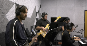 Four musicians play together in a soundproof studio. One person in the foreground plays an electric guitar, another plays a bass guitar, a third plays keyboard and sings into a microphone, and the fourth plays another electric guitar. All are wearing headphones and appear focused on their instruments. The room has gray acoustic panels on the walls and sheet music stands in front of some of the musicians.