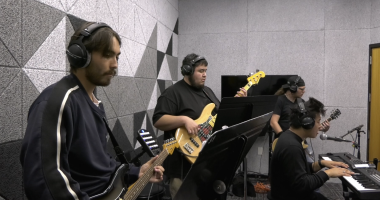 Four musicians play together in a soundproof studio. One person in the foreground plays an electric guitar, another plays a bass guitar, a third plays keyboard and sings into a microphone, and the fourth plays another electric guitar. All are wearing headphones and appear focused on their instruments. The room has gray acoustic panels on the walls and sheet music stands in front of some of the musicians.