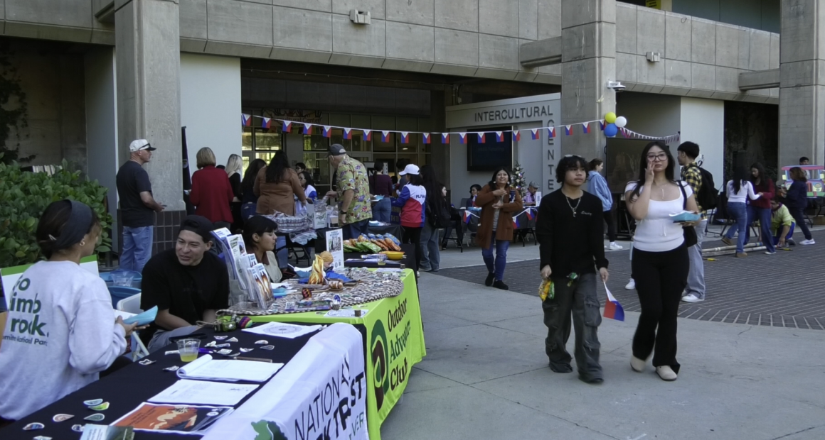 A lively outdoor event takes place in front of a building labeled “Intercultural Center.” Several tables with banners and displays are set up, including one for the “Outdoor Adventure Club” and “National Park Trust.” People are interacting at booths, walking around, and enjoying food. Colorful flags and balloons decorate the area, and the atmosphere appears casual and social.