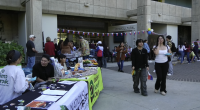 A lively outdoor event takes place in front of a building labeled “Intercultural Center.” Several tables with banners and displays are set up, including one for the “Outdoor Adventure Club” and “National Park Trust.” People are interacting at booths, walking around, and enjoying food. Colorful flags and balloons decorate the area, and the atmosphere appears casual and social.