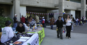 A lively outdoor event takes place in front of a building labeled “Intercultural Center.” Several tables with banners and displays are set up, including one for the “Outdoor Adventure Club” and “National Park Trust.” People are interacting at booths, walking around, and enjoying food. Colorful flags and balloons decorate the area, and the atmosphere appears casual and social.