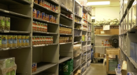 Storage room with shelves stocked with canned goods, boxes, and plastic bins, resembling a food pantry or supply room with fluorescent lighting overhead.