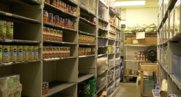 Storage room with shelves stocked with canned goods, boxes, and plastic bins, resembling a food pantry or supply room with fluorescent lighting overhead.