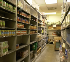 Storage room with shelves stocked with canned goods, boxes, and plastic bins, resembling a food pantry or supply room with fluorescent lighting overhead.