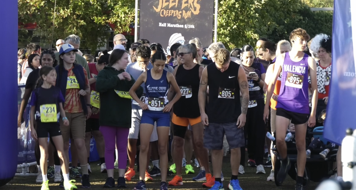 A group of runners stands at the starting line of a race called the “Jeepers Creepers Run,” as indicated by the banner in the background that also mentions “Half Marathon 5/10k.” Participants of various ages wear numbered race bibs and athletic attire, with some in team uniforms like “Fillmore Cross Country” and “Valencia.” The crowd behind them appears energetic and ready for the event, with trees and sunlight in the background.