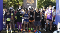 A group of runners stands at the starting line of a race called the “Jeepers Creepers Run,” as indicated by the banner in the background that also mentions “Half Marathon 5/10k.” Participants of various ages wear numbered race bibs and athletic attire, with some in team uniforms like “Fillmore Cross Country” and “Valencia.” The crowd behind them appears energetic and ready for the event, with trees and sunlight in the background.