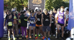 A group of runners stands at the starting line of a race called the “Jeepers Creepers Run,” as indicated by the banner in the background that also mentions “Half Marathon 5/10k.” Participants of various ages wear numbered race bibs and athletic attire, with some in team uniforms like “Fillmore Cross Country” and “Valencia.” The crowd behind them appears energetic and ready for the event, with trees and sunlight in the background.