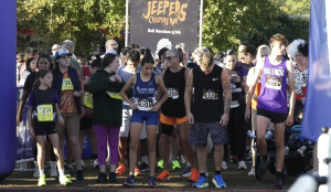 A group of runners stands at the starting line of a race called the “Jeepers Creepers Run,” as indicated by the banner in the background that also mentions “Half Marathon 5/10k.” Participants of various ages wear numbered race bibs and athletic attire, with some in team uniforms like “Fillmore Cross Country” and “Valencia.” The crowd behind them appears energetic and ready for the event, with trees and sunlight in the background.