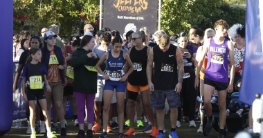 A group of runners stands at the starting line of a race called the “Jeepers Creepers Run,” as indicated by the banner in the background that also mentions “Half Marathon 5/10k.” Participants of various ages wear numbered race bibs and athletic attire, with some in team uniforms like “Fillmore Cross Country” and “Valencia.” The crowd behind them appears energetic and ready for the event, with trees and sunlight in the background.