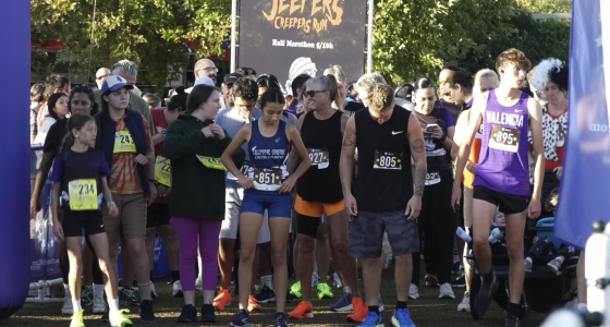 A group of runners stands at the starting line of a race called the “Jeepers Creepers Run,” as indicated by the banner in the background that also mentions “Half Marathon 5/10k.” Participants of various ages wear numbered race bibs and athletic attire, with some in team uniforms like “Fillmore Cross Country” and “Valencia.” The crowd behind them appears energetic and ready for the event, with trees and sunlight in the background.