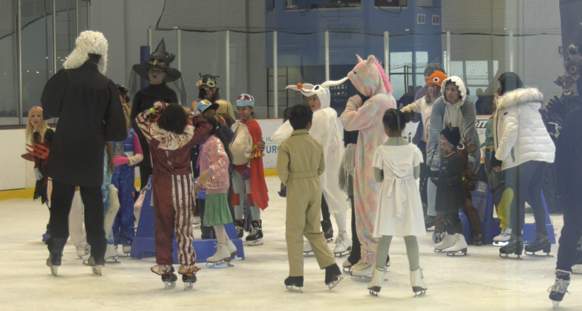 A group of children and adults in Halloween costumes ice skating at an indoor rink. Costumes include a unicorn, a witch, a ghost, a deer, a person in a judge’s robe with a white wig, and various colorful outfits. The group appears to be gathered in the middle of the rink, chatting and having fun.