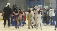 A group of children and adults in Halloween costumes ice skating at an indoor rink. Costumes include a unicorn, a witch, a ghost, a deer, a person in a judge’s robe with a white wig, and various colorful outfits. The group appears to be gathered in the middle of the rink, chatting and having fun.