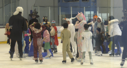 A group of children and adults in Halloween costumes ice skating at an indoor rink. Costumes include a unicorn, a witch, a ghost, a deer, a person in a judge’s robe with a white wig, and various colorful outfits. The group appears to be gathered in the middle of the rink, chatting and having fun.