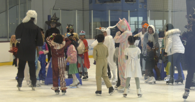 A group of children and adults in Halloween costumes ice skating at an indoor rink. Costumes include a unicorn, a witch, a ghost, a deer, a person in a judge’s robe with a white wig, and various colorful outfits. The group appears to be gathered in the middle of the rink, chatting and having fun.