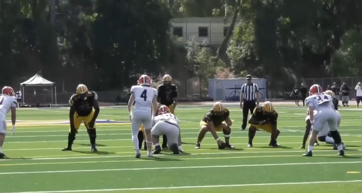 Alt text: A football game scene showing the offensive line of a team in black jerseys and yellow pants lined up against defenders in white uniforms with red helmets. The players are set before the snap on a sunny day, with a referee and spectators visible in the background on the field.