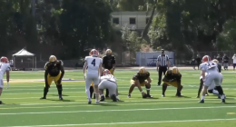 Alt text: A football game scene showing the offensive line of a team in black jerseys and yellow pants lined up against defenders in white uniforms with red helmets. The players are set before the snap on a sunny day, with a referee and spectators visible in the background on the field.