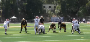 Alt text: A football game scene showing the offensive line of a team in black jerseys and yellow pants lined up against defenders in white uniforms with red helmets. The players are set before the snap on a sunny day, with a referee and spectators visible in the background on the field.