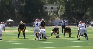 Alt text: A football game scene showing the offensive line of a team in black jerseys and yellow pants lined up against defenders in white uniforms with red helmets. The players are set before the snap on a sunny day, with a referee and spectators visible in the background on the field.