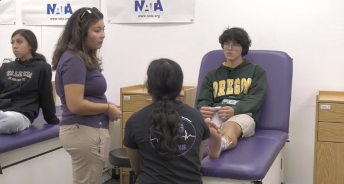 A student wearing an Oregon hoodie sits on a medical table with an ice pack wrapped around their ankle, while two people—one standing and one kneeling—appear to be discussing or examining the injury. Another student sits on a nearby table observing. The setting looks like a school athletic training or sports medicine room, with NATA banners on the wall.