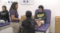 A student wearing an Oregon hoodie sits on a medical table with an ice pack wrapped around their ankle, while two people—one standing and one kneeling—appear to be discussing or examining the injury. Another student sits on a nearby table observing. The setting looks like a school athletic training or sports medicine room, with NATA banners on the wall.
