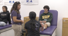 A student wearing an Oregon hoodie sits on a medical table with an ice pack wrapped around their ankle, while two people—one standing and one kneeling—appear to be discussing or examining the injury. Another student sits on a nearby table observing. The setting looks like a school athletic training or sports medicine room, with NATA banners on the wall.