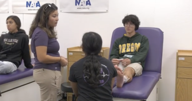 A student wearing an Oregon hoodie sits on a medical table with an ice pack wrapped around their ankle, while two people—one standing and one kneeling—appear to be discussing or examining the injury. Another student sits on a nearby table observing. The setting looks like a school athletic training or sports medicine room, with NATA banners on the wall.