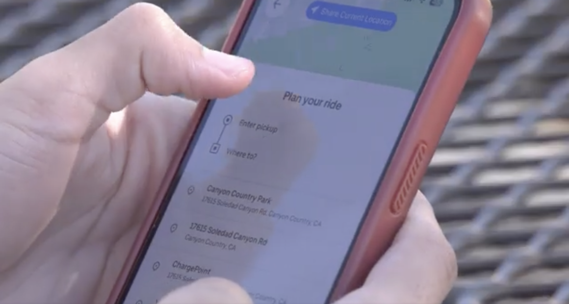 A person holds a smartphone with a red case, using a ride-planning app that displays “Plan your ride” and location options such as “Canyon Country Park” and “17615 Soledad Canyon Rd” on the screen.