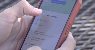 A person holds a smartphone with a red case, using a ride-planning app that displays “Plan your ride” and location options such as “Canyon Country Park” and “17615 Soledad Canyon Rd” on the screen.